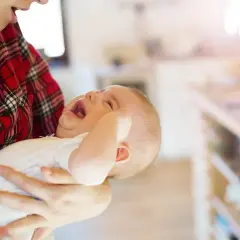 Płaczliwe niemowlę objawy: maluch w białej koszulce, z otwartymi ustami, trzymany przez mamę w czerwonej koszuli w kratę.