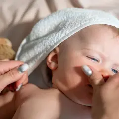 Delikatna pielęgnacja noworodka: mama delikatnie dotyka policzka dziecka w białym ręczniku, obok leży szczotka.
