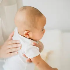 Maluch w białej koszulce, trzymany przez mamę. Czy po karmieniu piersią trzeba odbić? To pytanie nurtuje wielu rodziców.