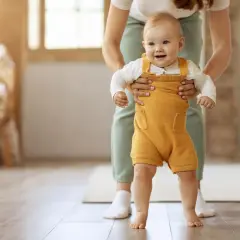 Uśmiechnięte dziecko w żółtym śpiochu stawia pierwsze kroki, wspierane przez rodzica. To piękny moment, kiedy dziecko zaczyna chodzić.