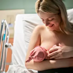 Uśmiechnięta mama karmi piersią noworodka. W szpitalu, obok łóżeczka, czeka na dziecko. Czasem karmienie może być trudne, np. gdy pojawi się grzybica sutka.