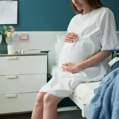 Kobieta w białej koszuli szpitalnej, z dłońmi na brzuchu, siedzi na łóżku. To już 3 trymestr, czas oczekiwania na dziecko.