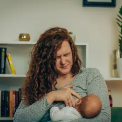 Kobieta z rudymi włosami karmi dziecko, jej twarz wykrzywiona bólem. Bolące sutki przy karmieniu to trudny początek macierzyństwa.