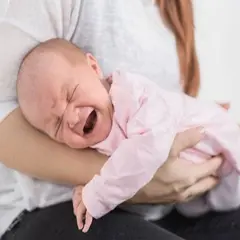 Noworodek w różowym ubranku płacze, cierpiąc z powodu bólu brzucha. Mama tuli go, próbując ukoić.