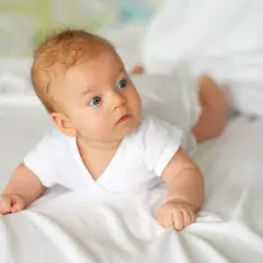 Niebieskooki 10 tygodniowe dziecko w białej koszulce leży na brzuchu na białej pościeli, badając otoczenie.
