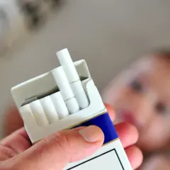 Dłoń trzyma paczkę papierosów na tle rozmazanego dziecka. Palenie a karmienie piersią to poważny problem.