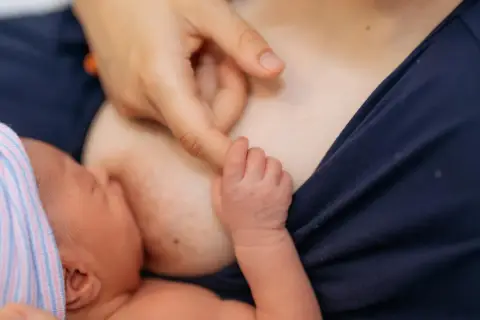 Nowe życie, pierwsze karmienie. Dłoń mamy delikatnie pieści główkę noworodka, który ssie mleko z piersi. Biały sutek po karmieniu świadczy o tym, że maluch jest najedzony.