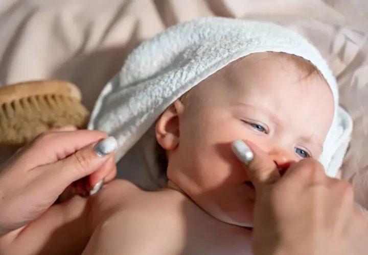 Delikatna pielęgnacja noworodka: mama delikatnie dotyka policzka dziecka w białym ręczniku, obok leży szczotka.