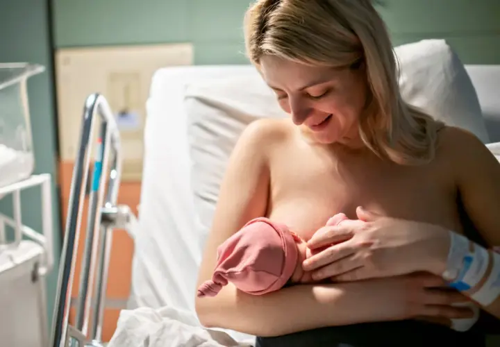 Uśmiechnięta mama karmi piersią noworodka. W szpitalu, obok łóżeczka, czeka na dziecko. Czasem karmienie może być trudne, np. gdy pojawi się grzybica sutka.