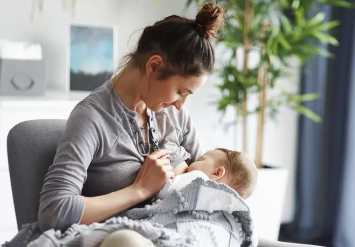 Kobieta karmi piersią dziecko, otulone kocykiem. Czy w ciąży można karmić piersią? To pytanie nurtuje wiele mam.