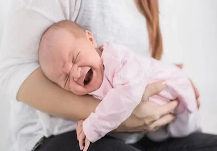 Noworodek w różowym ubranku płacze, cierpiąc z powodu bólu brzucha. Mama tuli go, próbując ukoić.