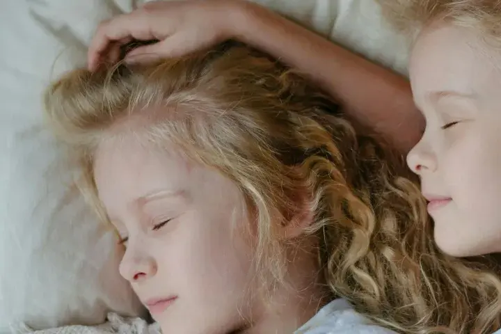 Dwie dziewczynki z blond włosami śpią blisko siebie. Jedna z nich ma rękę na głowie, co może sugerować niespokojny sen dwulatka.