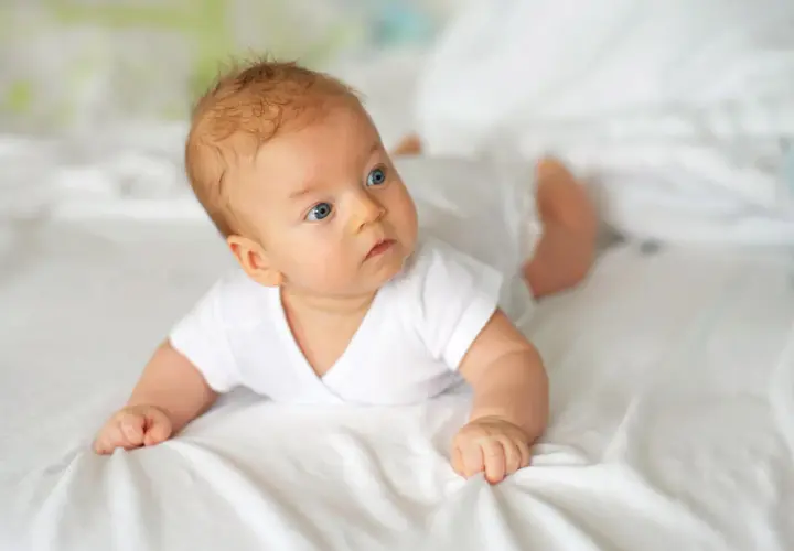 Niebieskooki 10 tygodniowe dziecko w białej koszulce leży na brzuchu na białej pościeli, badając otoczenie.
