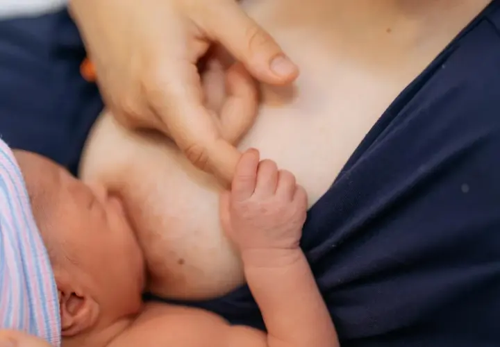 Nowe życie, pierwsze karmienie. Dłoń mamy delikatnie pieści główkę noworodka, który ssie mleko z piersi. Biały sutek po karmieniu świadczy o tym, że maluch jest najedzony.