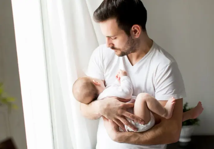 Ojciec tuli śpiące dziecko, które ma 24 tydzień życia. Delikatne światło wpada przez okno.