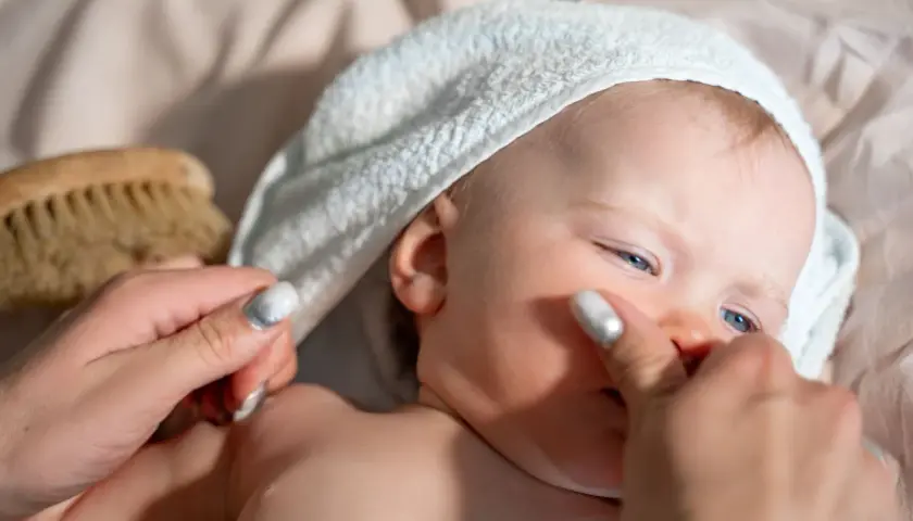 Delikatna pielęgnacja noworodka: mama delikatnie dotyka policzka dziecka w białym ręczniku, obok leży szczotka.