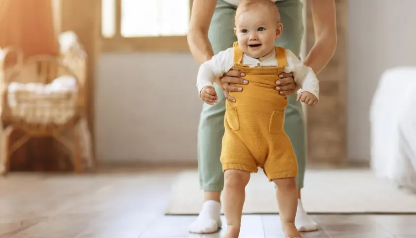 Uśmiechnięte dziecko w żółtym śpiochu stawia pierwsze kroki, wspierane przez rodzica. To piękny moment, kiedy dziecko zaczyna chodzić.