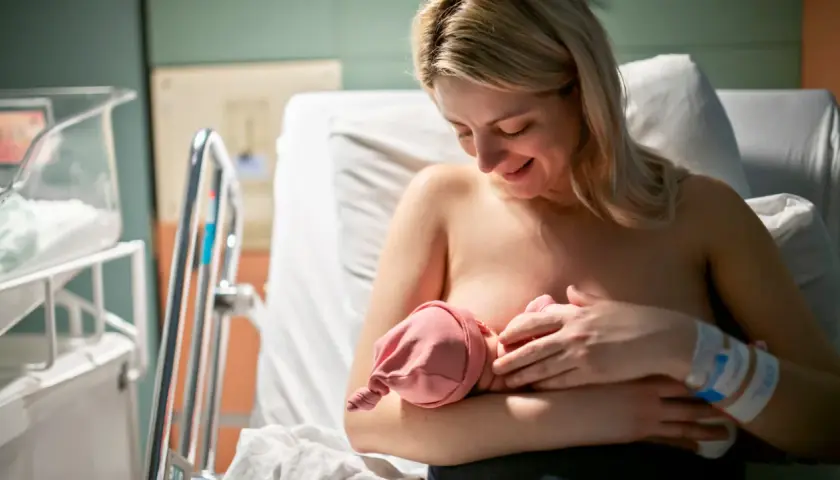 Uśmiechnięta mama karmi piersią noworodka. W szpitalu, obok łóżeczka, czeka na dziecko. Czasem karmienie może być trudne, np. gdy pojawi się grzybica sutka.
