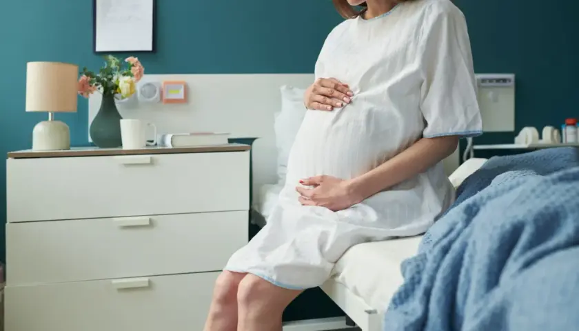 Kobieta w białej koszuli szpitalnej, z dłońmi na brzuchu, siedzi na łóżku. To już 3 trymestr, czas oczekiwania na dziecko.