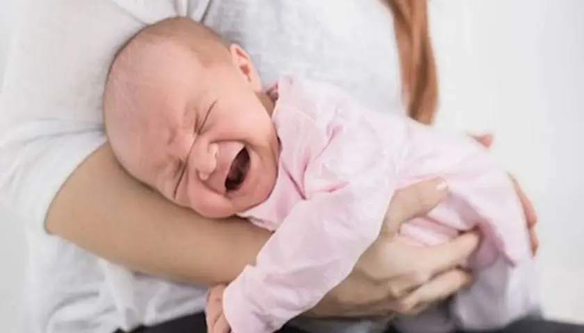 Noworodek w różowym ubranku płacze, cierpiąc z powodu bólu brzucha. Mama tuli go, próbując ukoić.