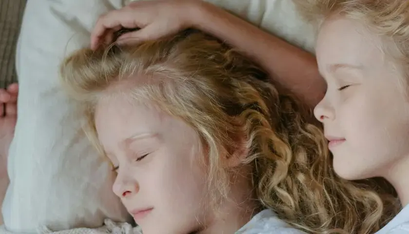 Dwie dziewczynki z blond włosami śpią blisko siebie. Jedna z nich ma rękę na głowie, co może sugerować niespokojny sen dwulatka.