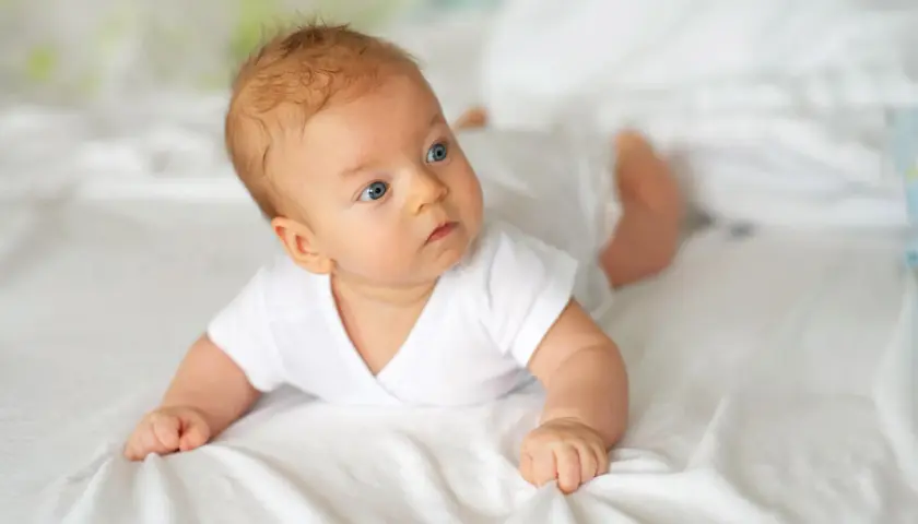 Niebieskooki 10 tygodniowe dziecko w białej koszulce leży na brzuchu na białej pościeli, badając otoczenie.