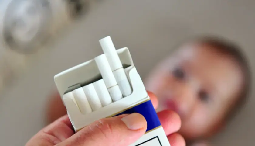 Dłoń trzyma paczkę papierosów na tle rozmazanego dziecka. Palenie a karmienie piersią to poważny problem.