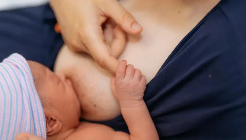 Nowe życie, pierwsze karmienie. Dłoń mamy delikatnie pieści główkę noworodka, który ssie mleko z piersi. Biały sutek po karmieniu świadczy o tym, że maluch jest najedzony.