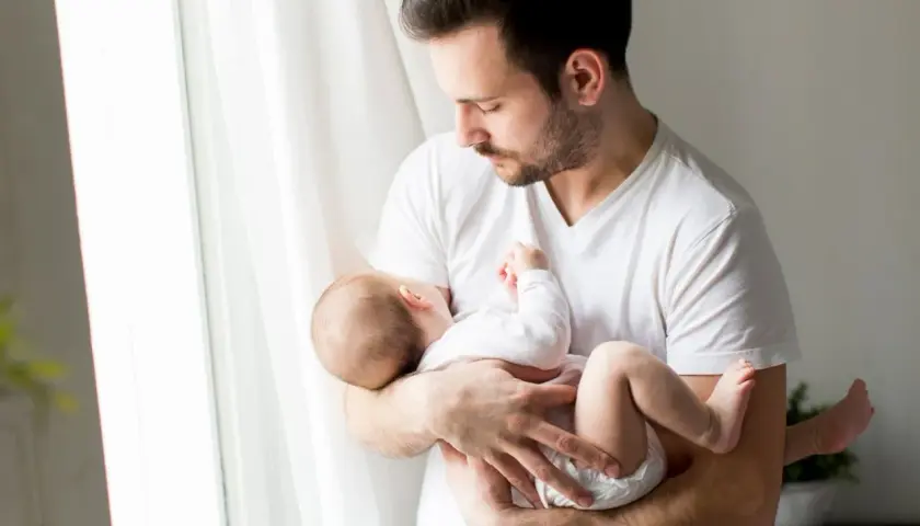 Ojciec tuli śpiące dziecko, które ma 24 tydzień życia. Delikatne światło wpada przez okno.