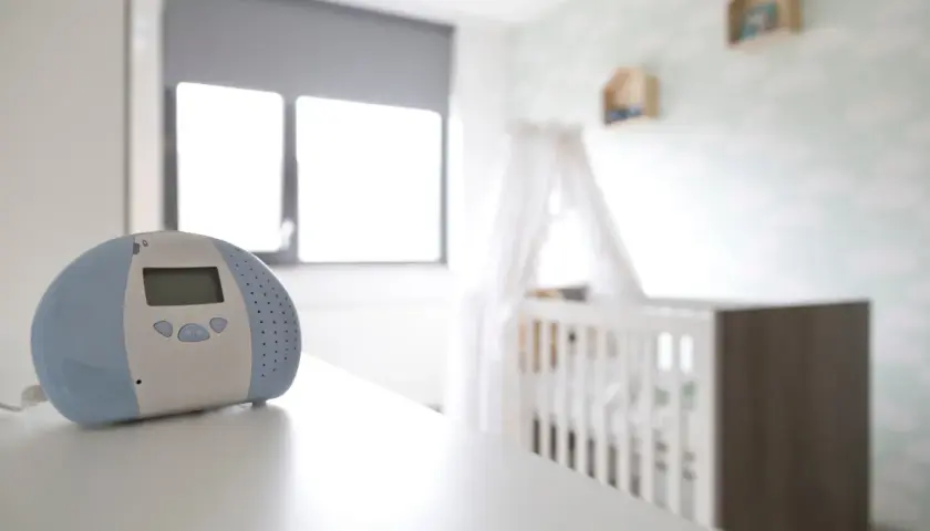 Niania elektroniczna czuwa, by usłyszeć każdy szumis dźwięk z pokoju dziecka. W tle białe łóżeczko i chmurki na ścianie.