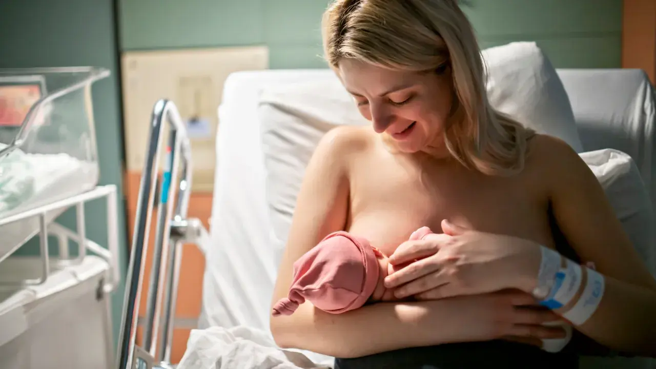 Uśmiechnięta mama karmi piersią noworodka. W szpitalu, obok łóżeczka, czeka na dziecko. Czasem karmienie może być trudne, np. gdy pojawi się grzybica sutka.