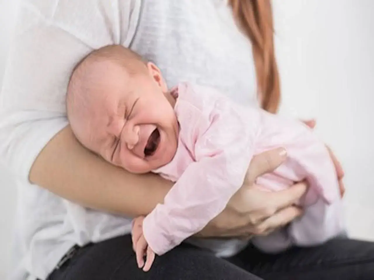 Noworodek w różowym ubranku płacze, cierpiąc z powodu bólu brzucha. Mama tuli go, próbując ukoić.