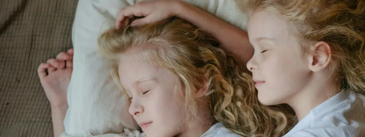 Dwie dziewczynki z blond włosami śpią blisko siebie. Jedna z nich ma rękę na głowie, co może sugerować niespokojny sen dwulatka.