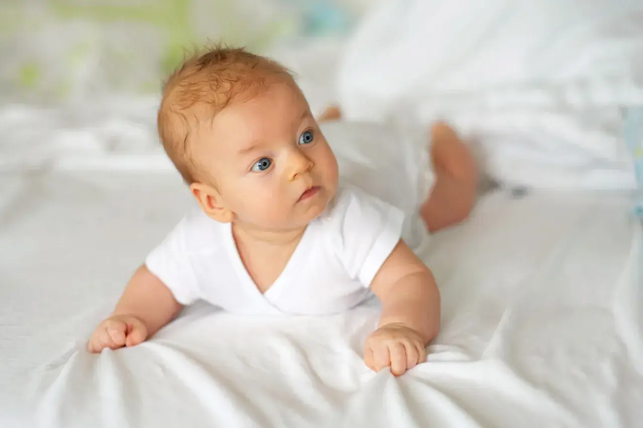 Niebieskooki 10 tygodniowe dziecko w białej koszulce leży na brzuchu na białej pościeli, badając otoczenie.