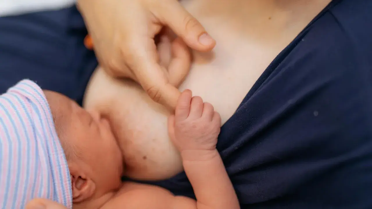Nowe życie, pierwsze karmienie. Dłoń mamy delikatnie pieści główkę noworodka, który ssie mleko z piersi. Biały sutek po karmieniu świadczy o tym, że maluch jest najedzony.
