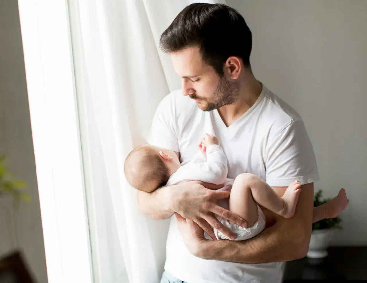 Ojciec tuli śpiące dziecko, które ma 24 tydzień życia. Delikatne światło wpada przez okno.