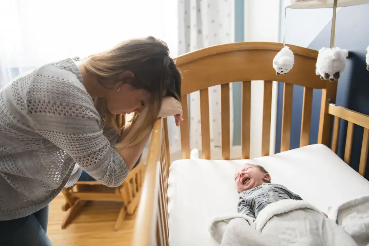 Zmęczona mama pochyla się nad ł&oacute;żeczkiem, gdzie płacze niemowlę. Czy to znak, że 7 miesięczne dziecko nie śpi wystarczająco?
