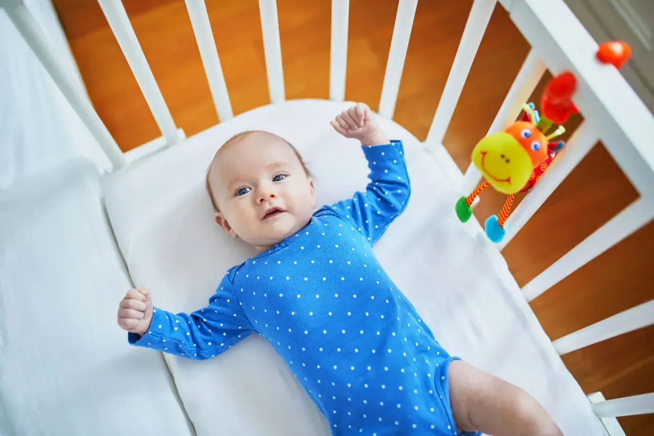 Urocze 10 tygodniowe dziecko w niebieskim bodziaku w kropki leży w łóżeczku, bawiąc się zabawką-żyrafką.